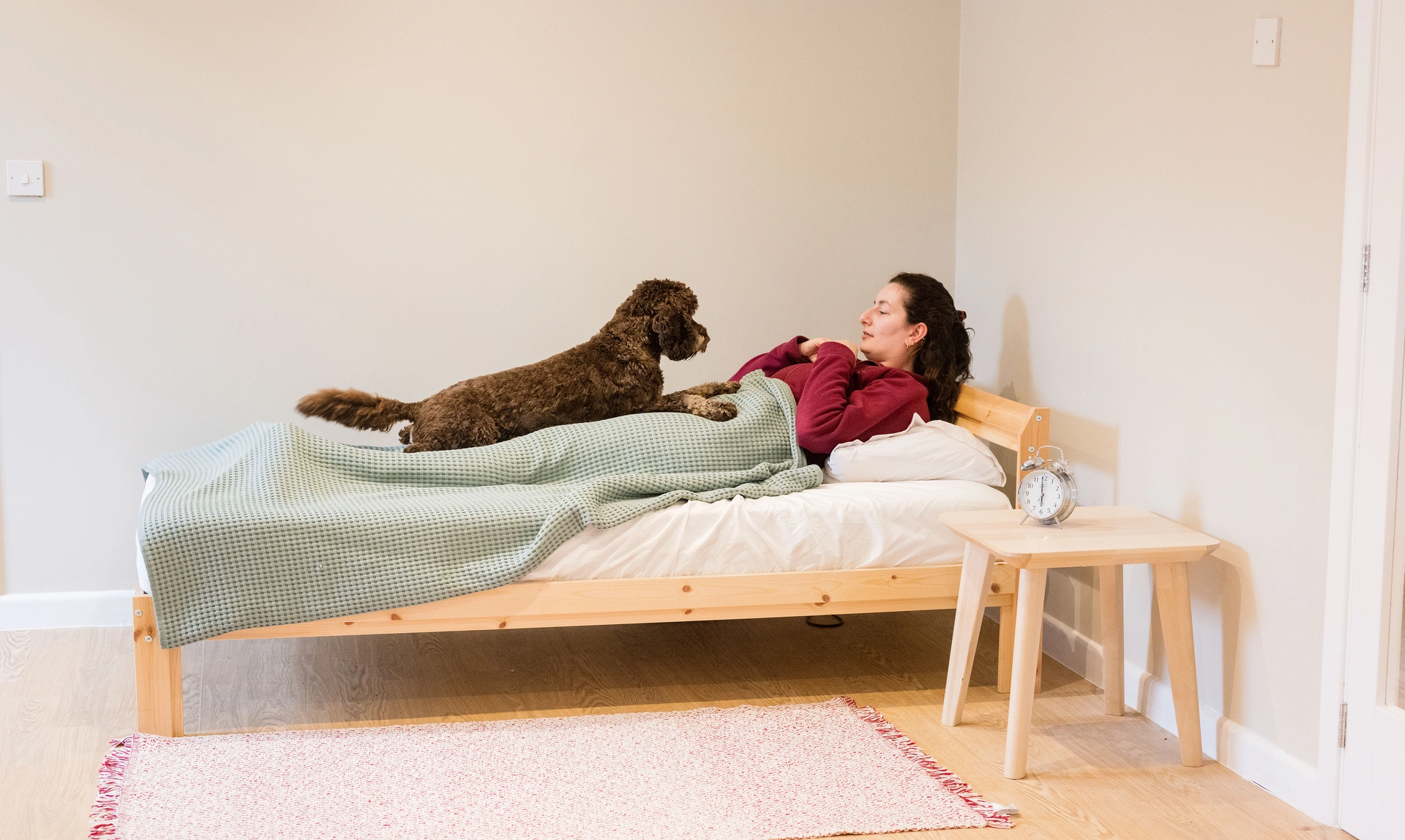 Woman in Hearing Dogs burgundy jacket in bed in room with a dog on the bed in front of her laying down but looking alertly at the women