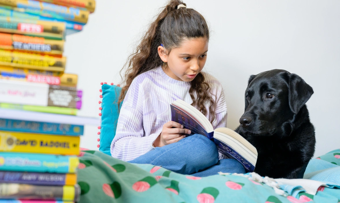 A young girl reads to a black labrador while they are at on a bed with a pile of books to their left of them