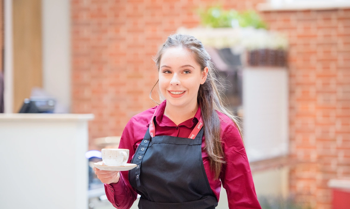 A young woman wearing a black apron and burgundy shirt holding a cup of coffee on a saucer