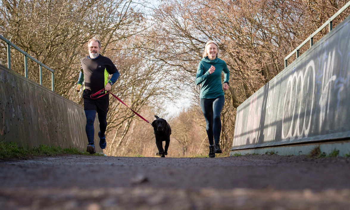 A man and a woman outside running along a bridge with a black labrador running between them