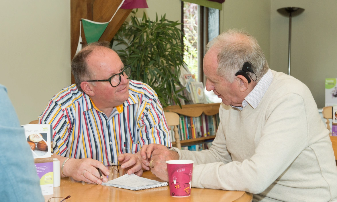 Two men sat at a table in a restaurant talking, one has a cochlear implant visible