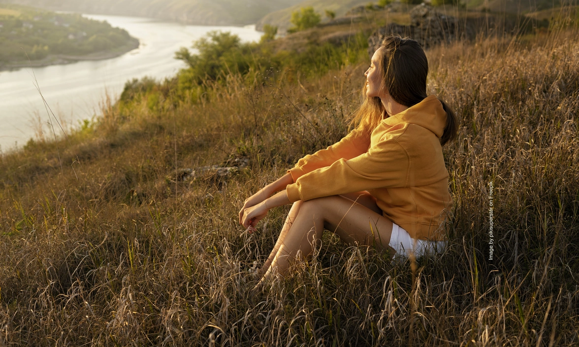 A woman sat on a hill as the sunsets looking out at some countryside
