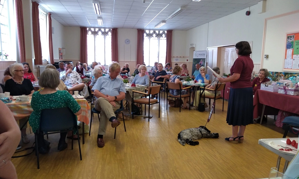 Woman in hearing dogs uniform speakers to a room full of people sat at tables with a sleeping dog at her feet