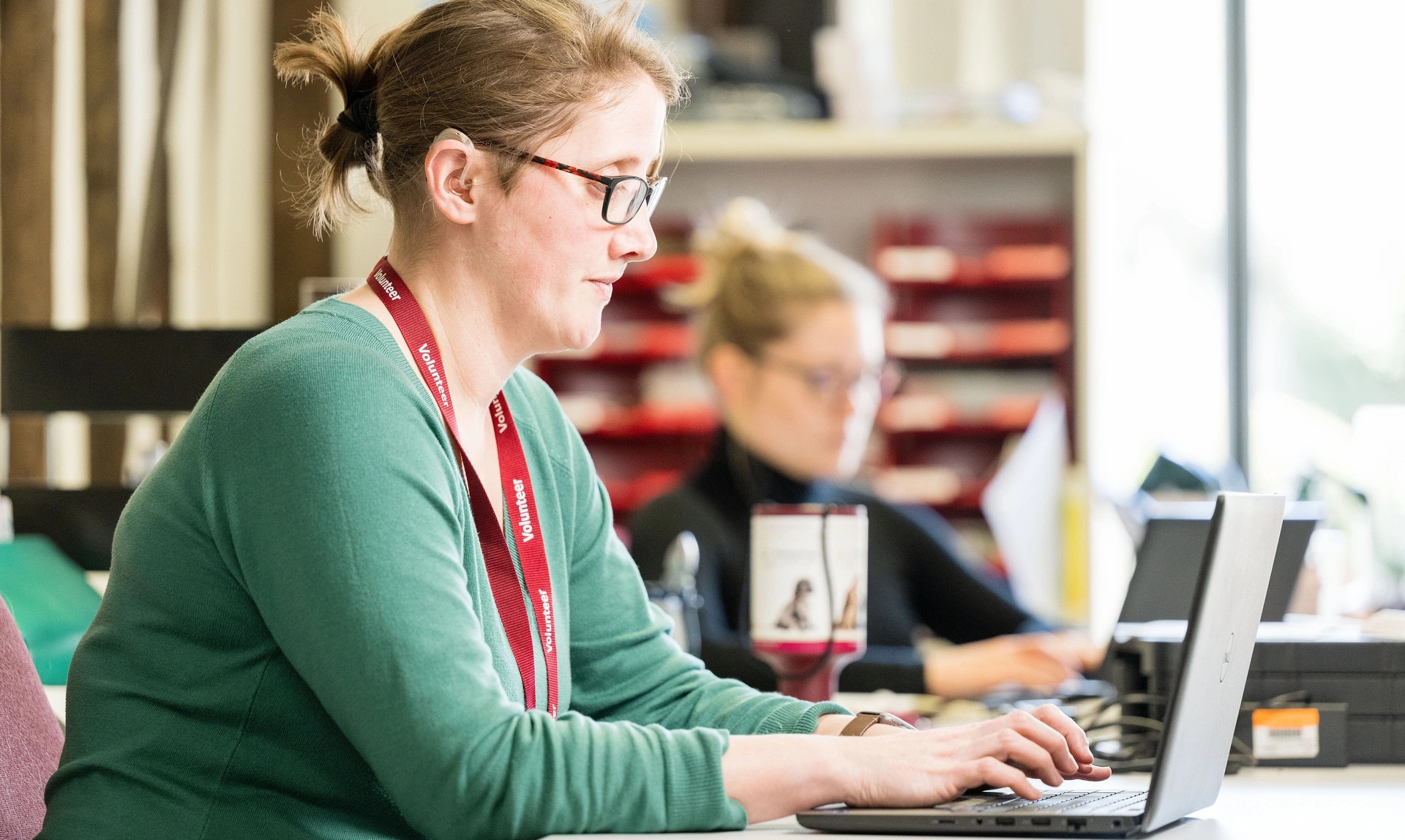 A woman in a green top with a burgundy hearing dogs landyard sat at a desk working on a laptop 