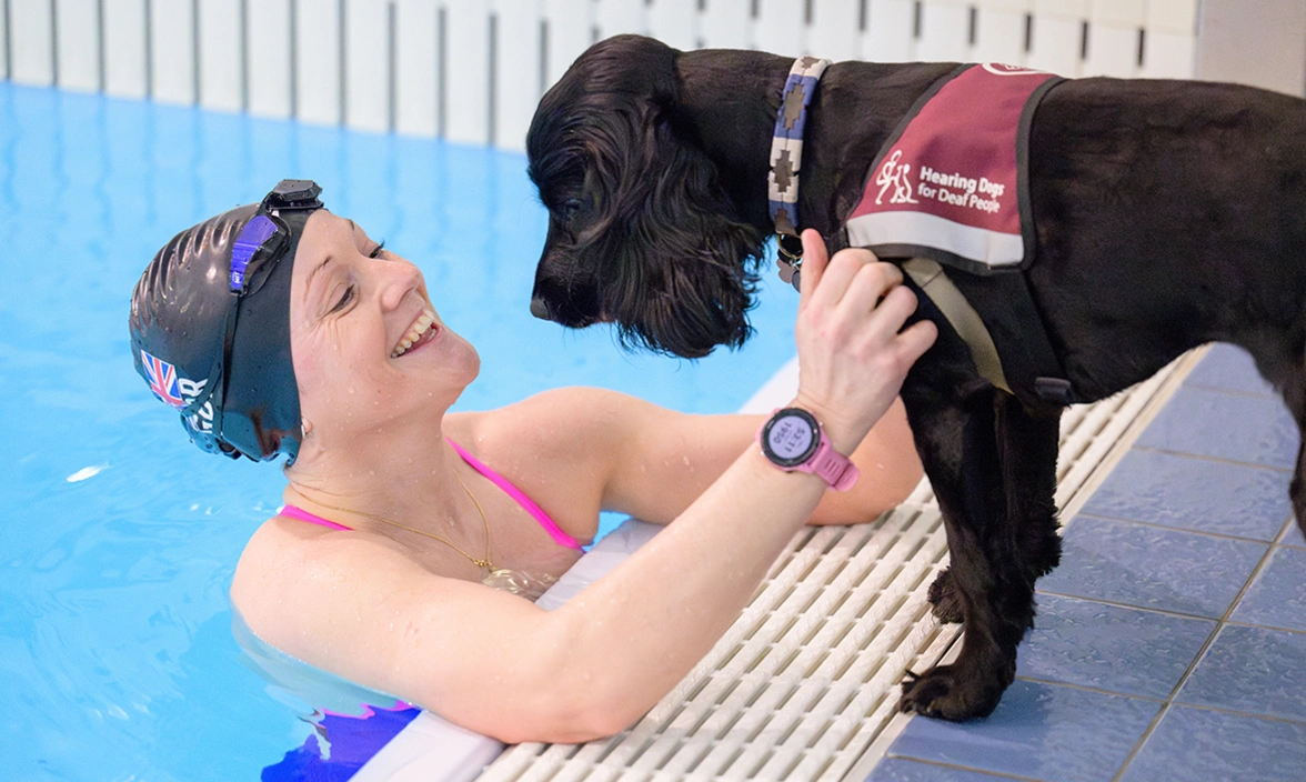 A woman in a swimming pool with a hearing dog in a jacket at the poolside looking at her