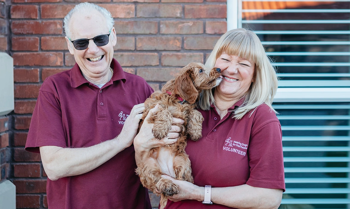 Two Hearing Dogs volunteers laughing and holding  a cockapoo puppy which is leaning towards the woman and trying to lick her face