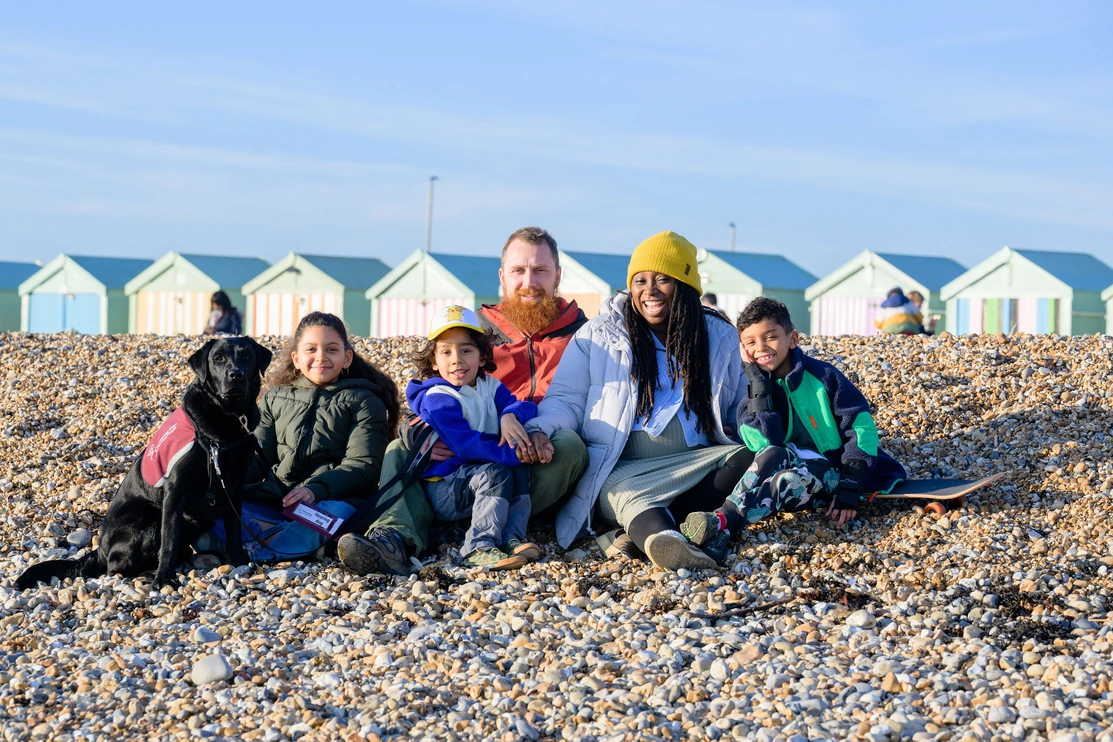 A family consisting of a dad, a mum, two younger boys and a slightly older girl with a black labrador are all sat on a pebble beach with beach huts in the background smiling at the camera