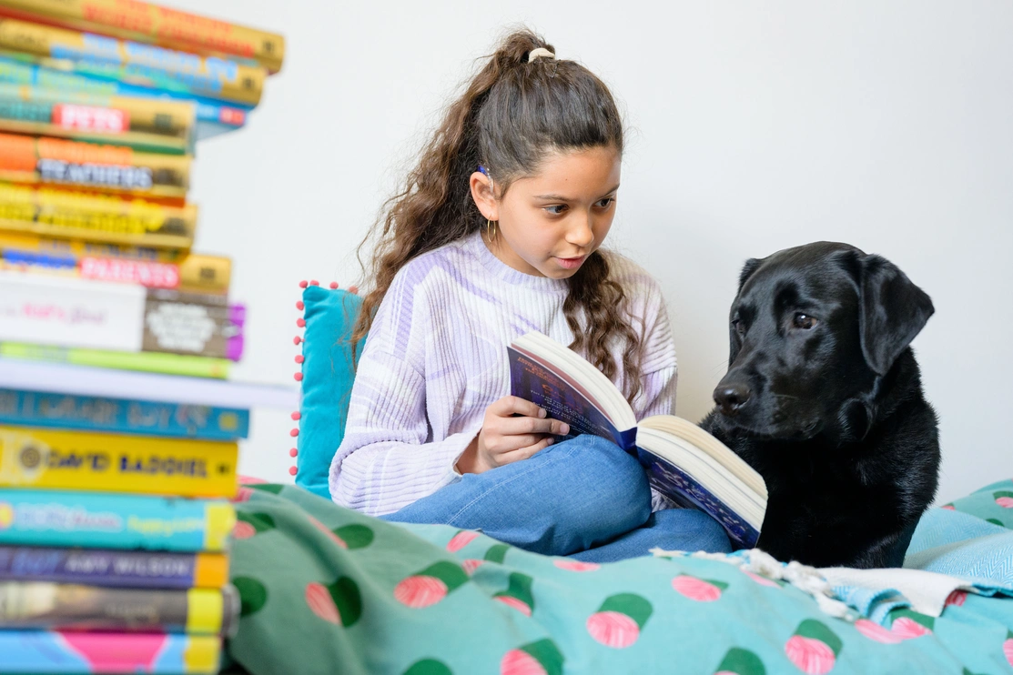 A young girl reads to a black labrador while they are at on a bed with a pile of books to their left of them