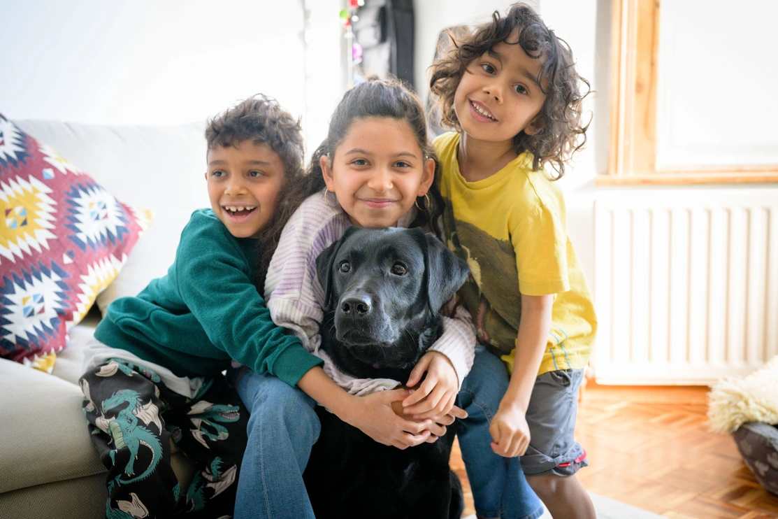 Two younger boys and a slightly older girl are smiling or laughing and huddled together all hugging a black labrador