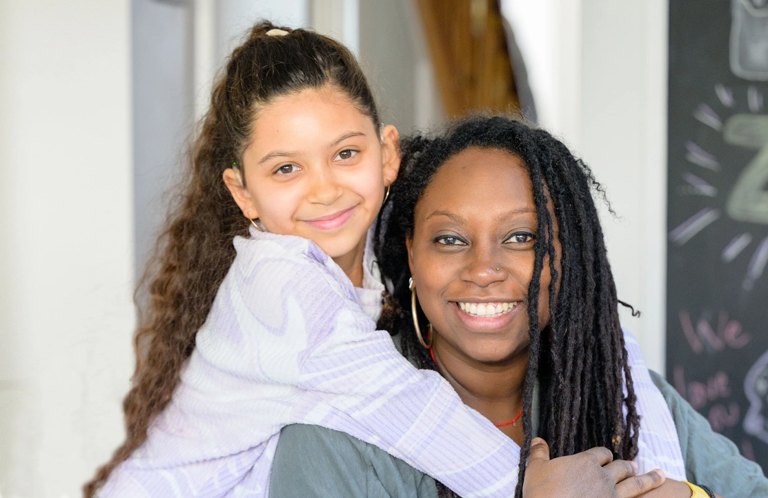 A young girl is stood up hugging her mum from behind, her mum is sat down both are smiling at the camera