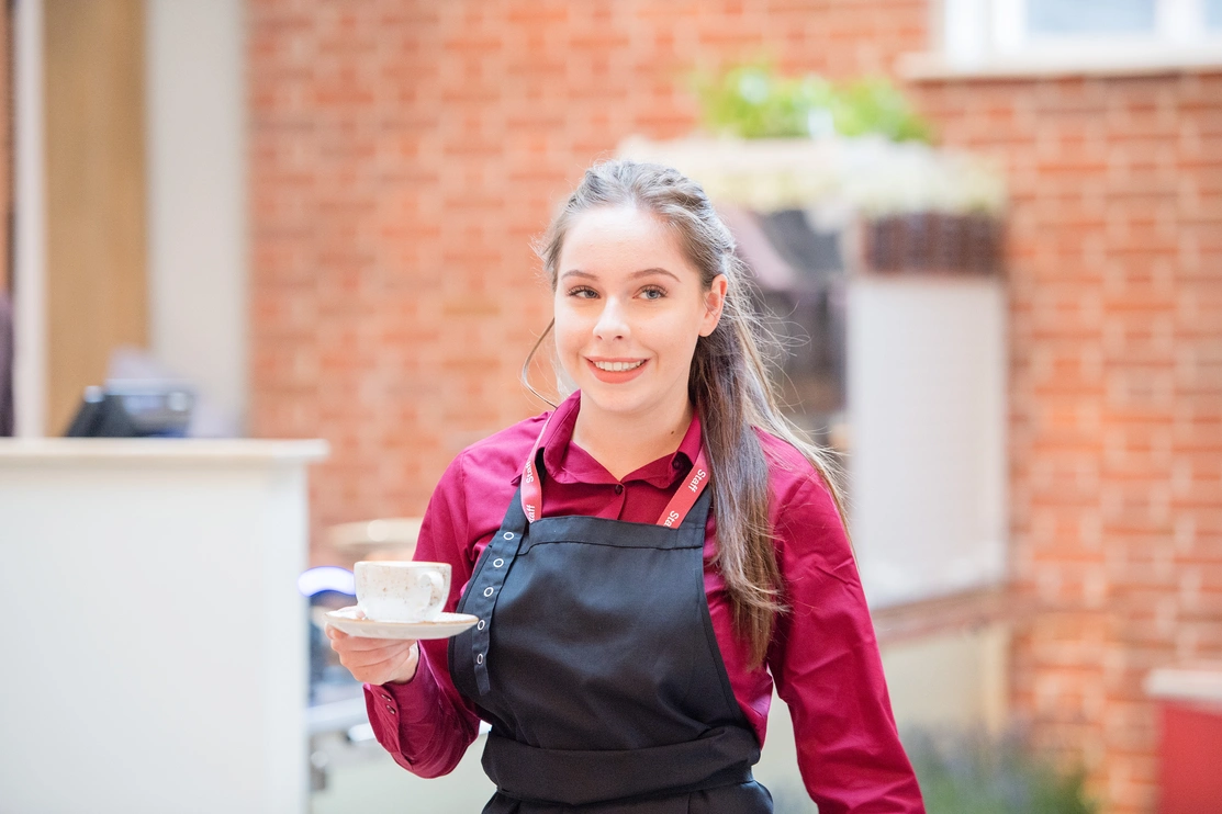 A young woman wearing a black apron and burgundy shirt holding a cup of coffee on a saucer