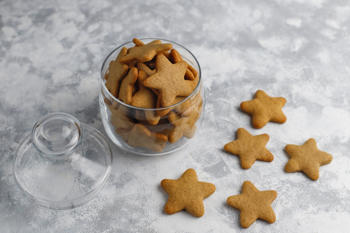 A glass jar filled with star-shaped gingerbread dog biscuits is open on a textured grey surface, with several biscuits scattered around it.