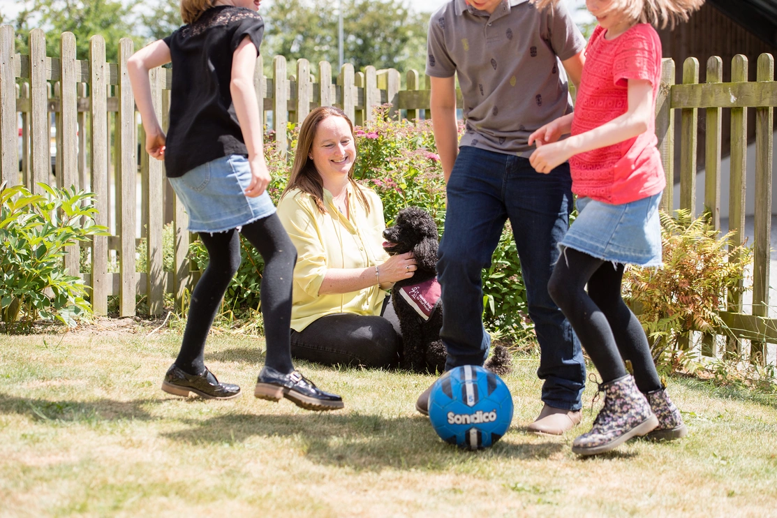 A woman is sat on grass in sunshine with her hearing dog poodle watching her three children playing football nearby
