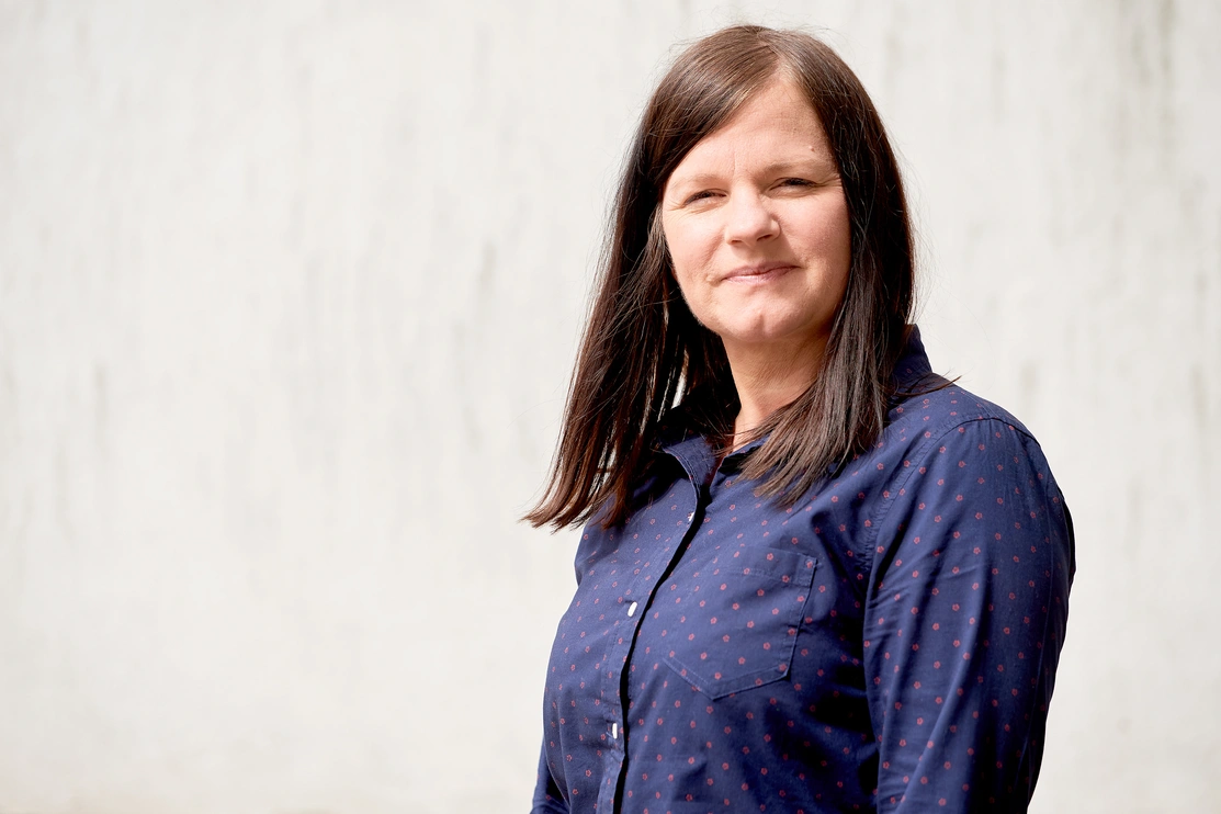 Mid shot of a woman with long brown hair and a dark blue shirt with a light grey textured background