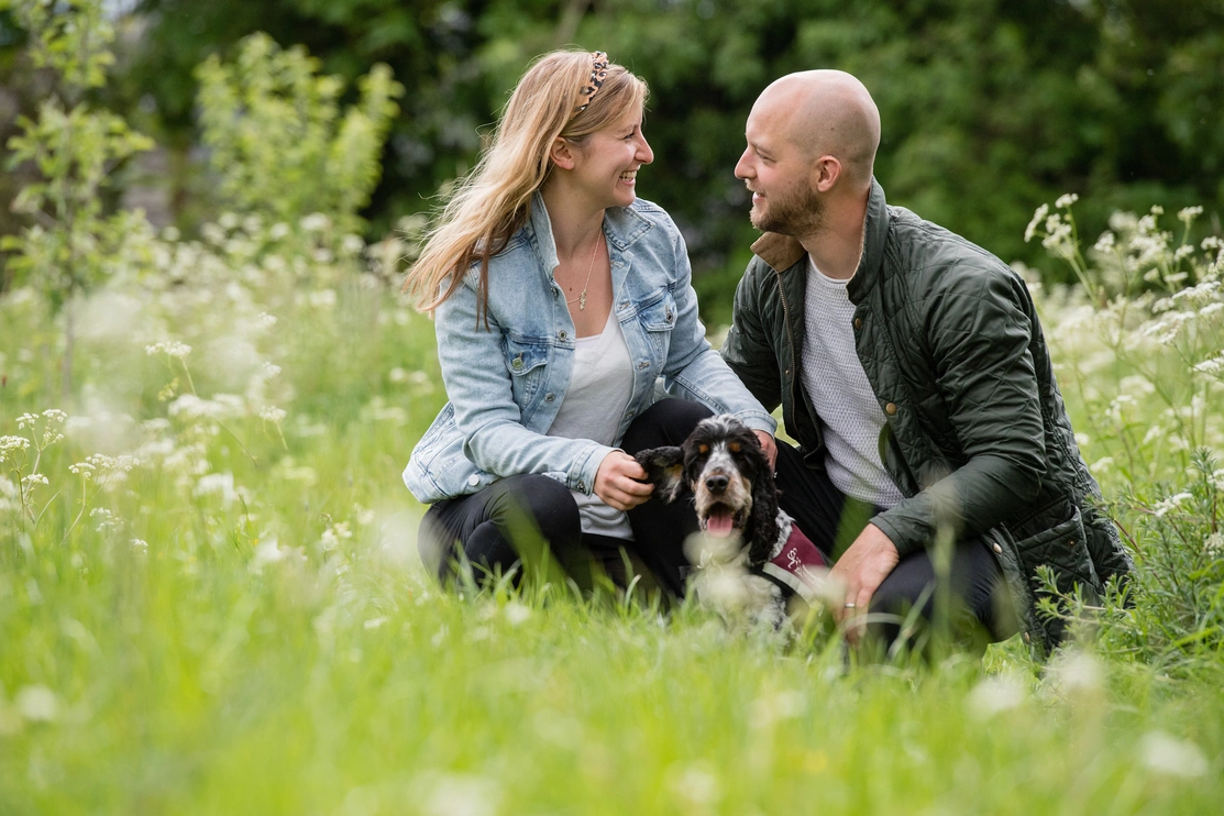 A man and a woman looking at each other in a field of flowers with a hearing dog Spaniel sat in front of them