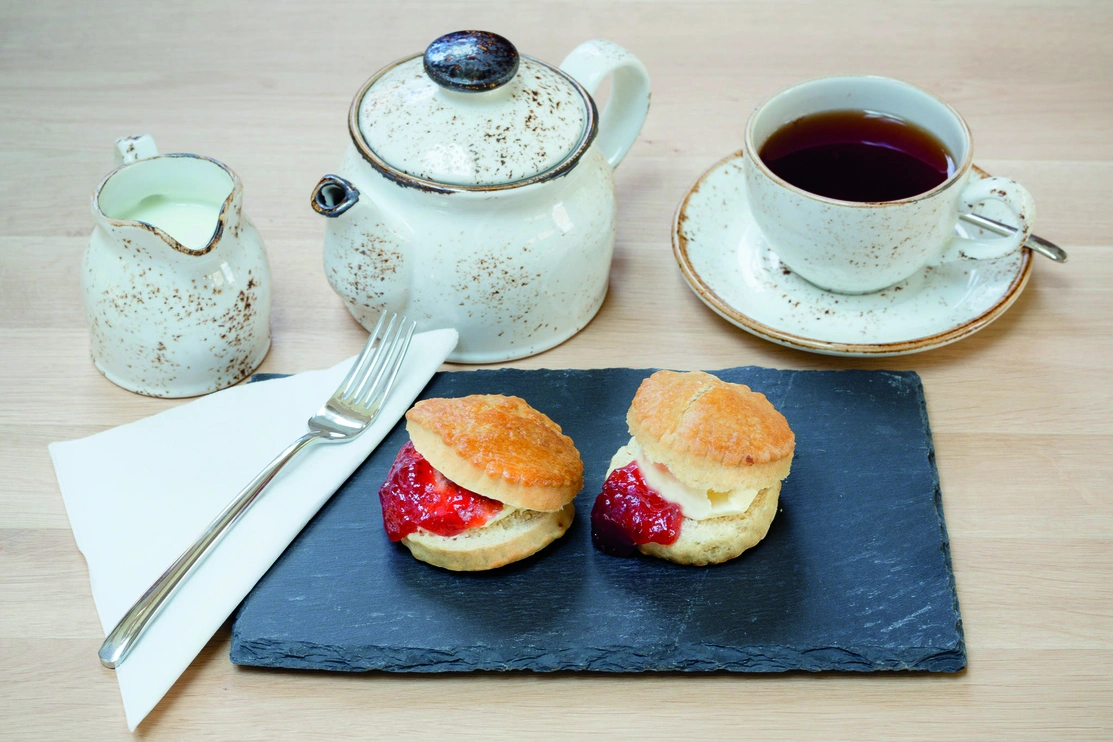 Teapot with cup of tea and milk, and two scones infront next to a fork and napkin