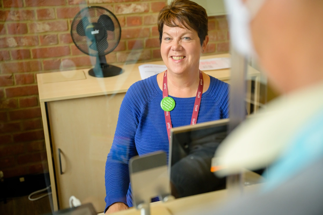 Smiling woman sat at a reception desk wearing a staff lanyard and a lipreading badge greeting a person