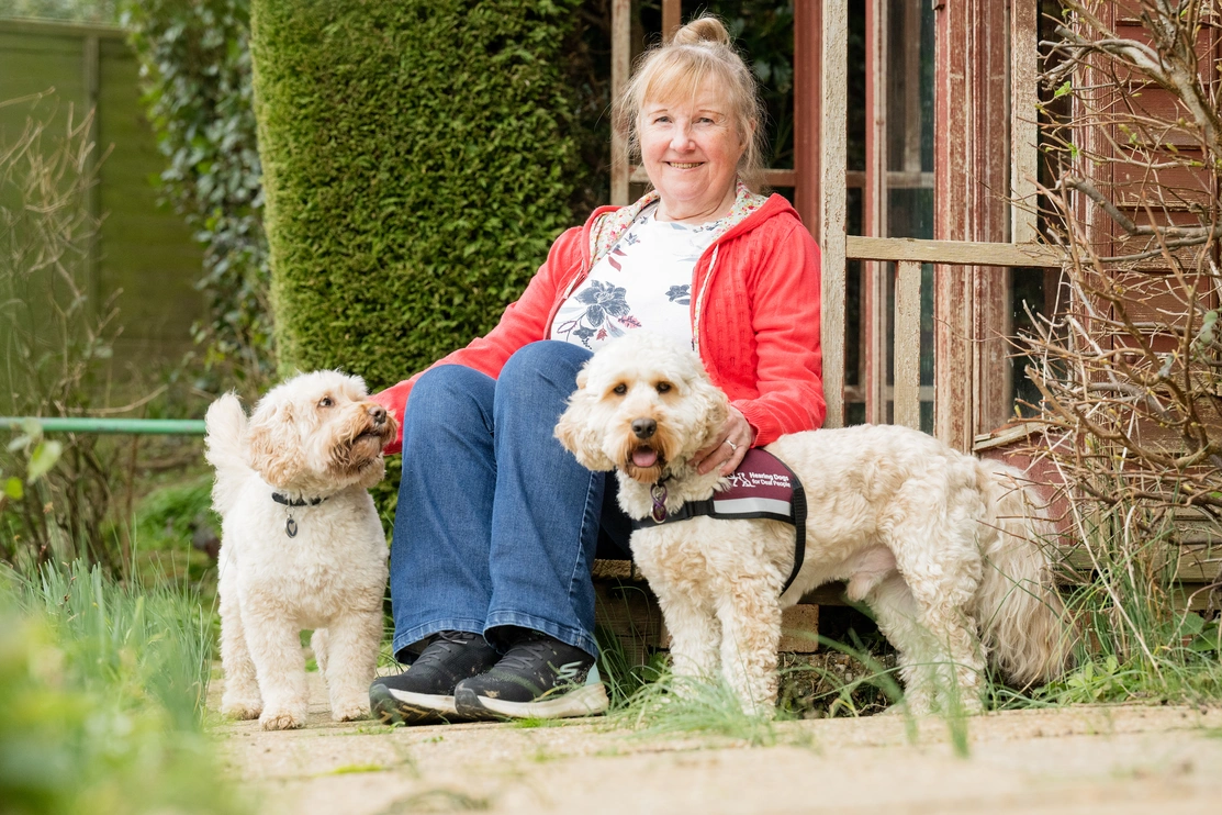 A woman stood in a garden smiling with two light coloured cockapoos, one if wearing a hearing dogs jacket