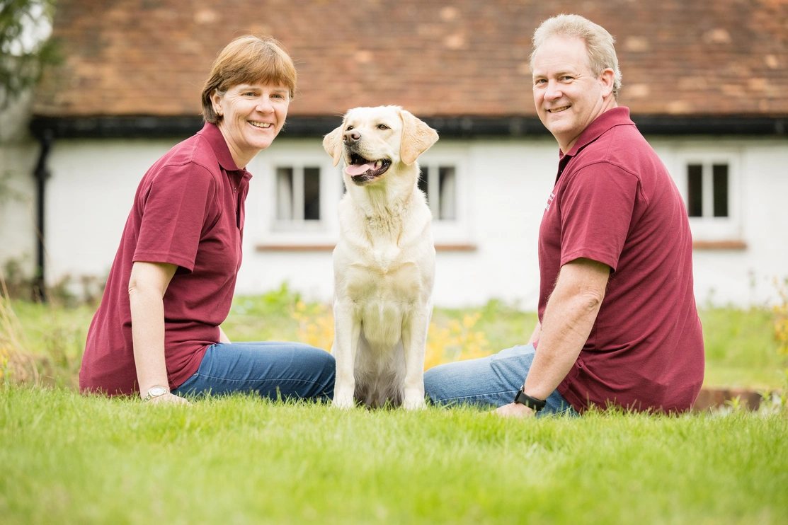A happy white labrador sitting on grass between a man and a woman in hearing dogs volunteer uniform.