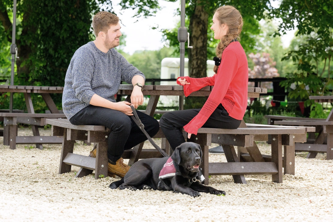 Two people sat on a bench outside chatting, one of them had a hearing dog wearing a qualified jacket
