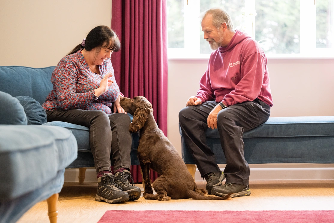 A woman is sat in a lounge with a man dressed in hearing dogs staff uniform meeting a brown spaniel