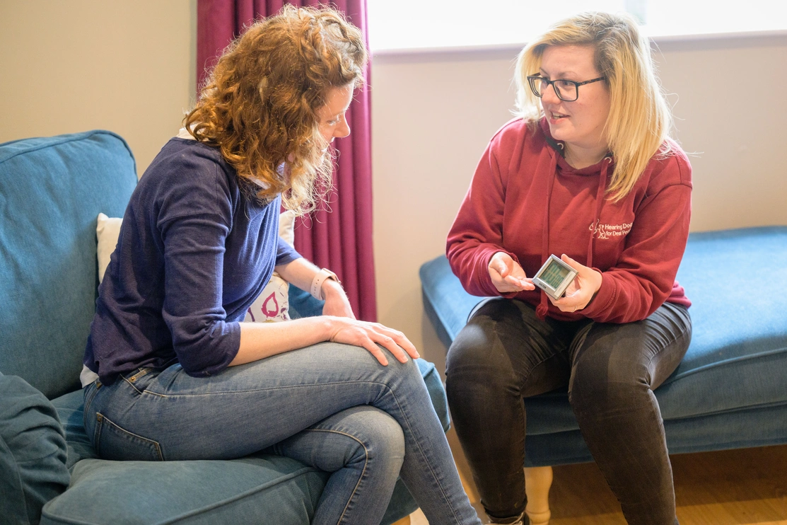 Two women sat in a lounge, the one is hearing dogs uniform is showing the other how to use a training timer