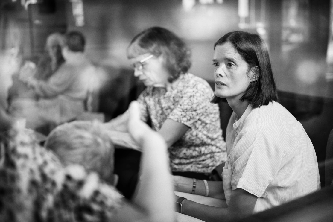 : A black and white image in a cafe setting, the focus is on a woman staring into the distance, looking worried and deep in thought, while conversations are happening around her