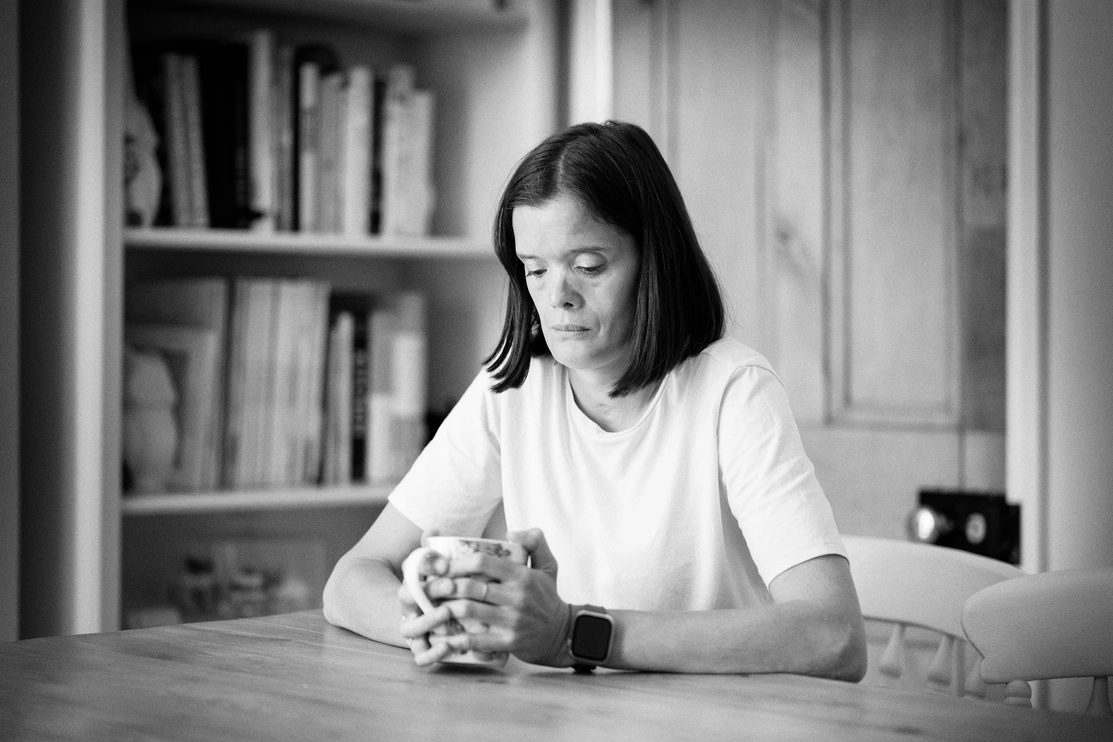 A black and white image of a woman sitting at a table looking down at a mug she is holding, she has an anxious and sad expression on her face