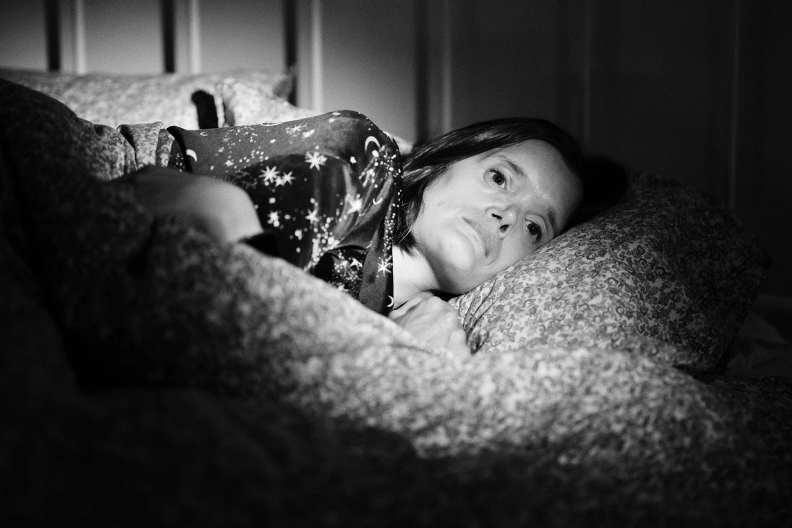 A black and white close-up image of a woman lying in bed, looking deep in thought, her eyes are open and she looks worried