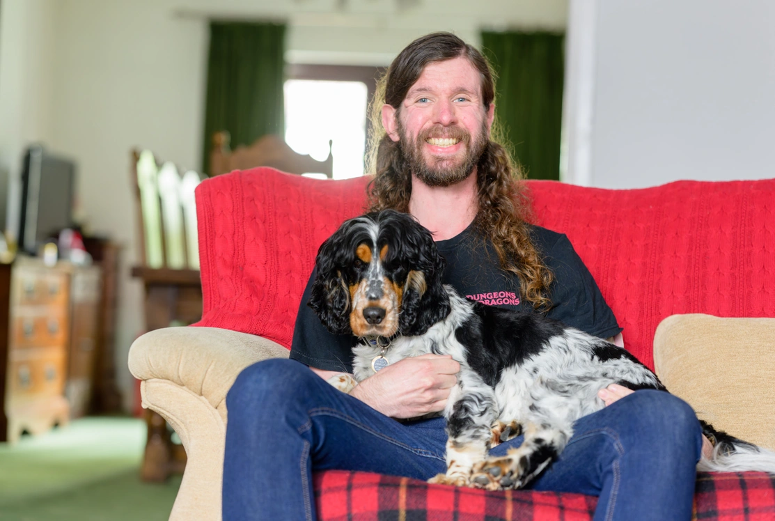 A young man with long brown hair is sat on a sofa with his arm around a black and white Spaniel, the man is laughing as he reads a book