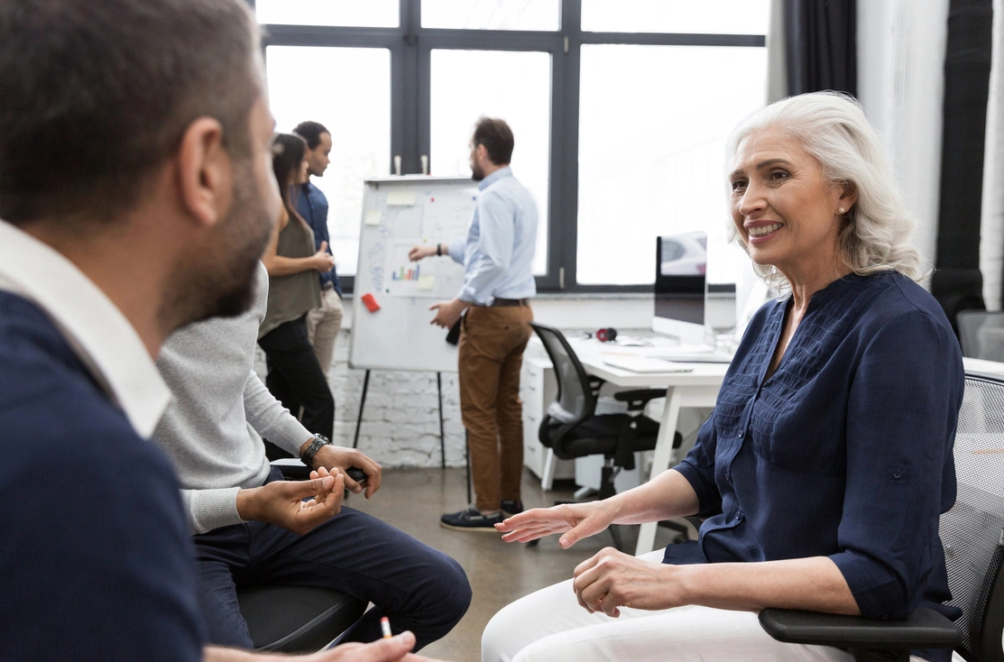 Three people sat talking in an open plan office with other people in the background