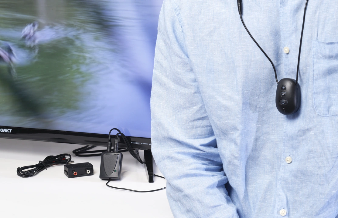 Close of of a person in a shirt wearing a hearing device around their neck with another device connected to the television behind them