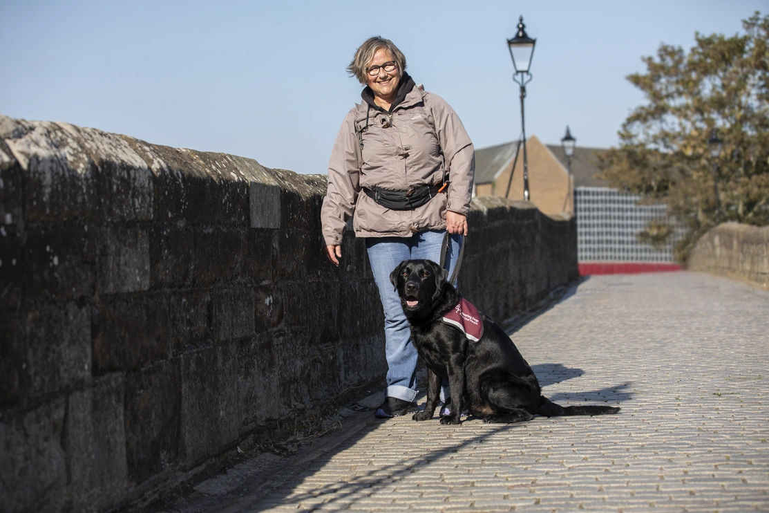 A woman standing next to a stone wall with a black labrador sitting at her feet by her side with a street light in the background