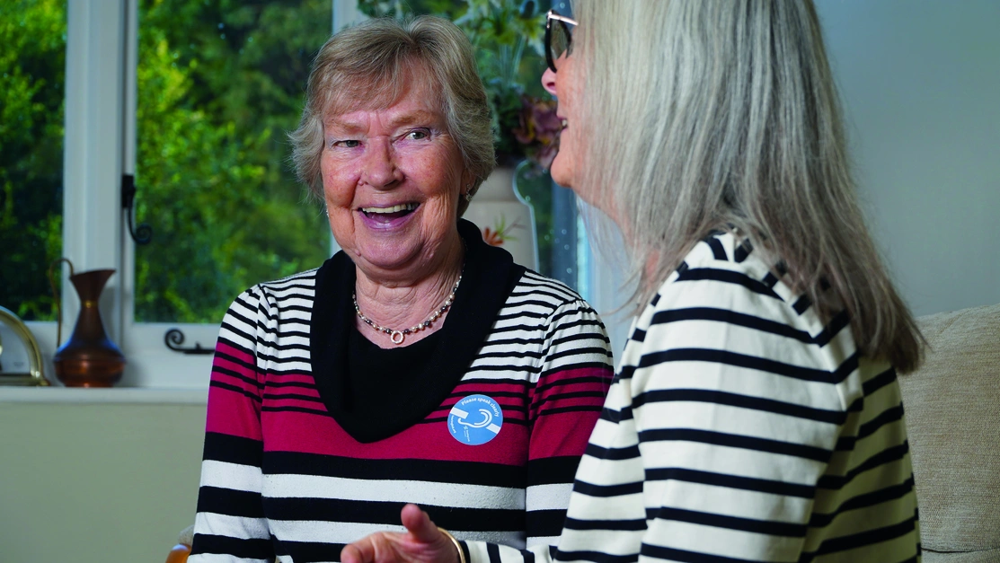Two older women talking and smiling, one if wearing a badge with a deaf symbol that says ‘Please speak clearly’