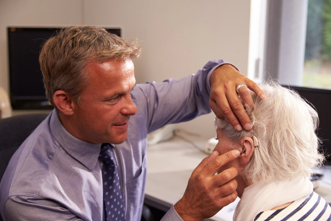 A male audiologist is fitting a hearing aid to the ear of a woman sat beside him
