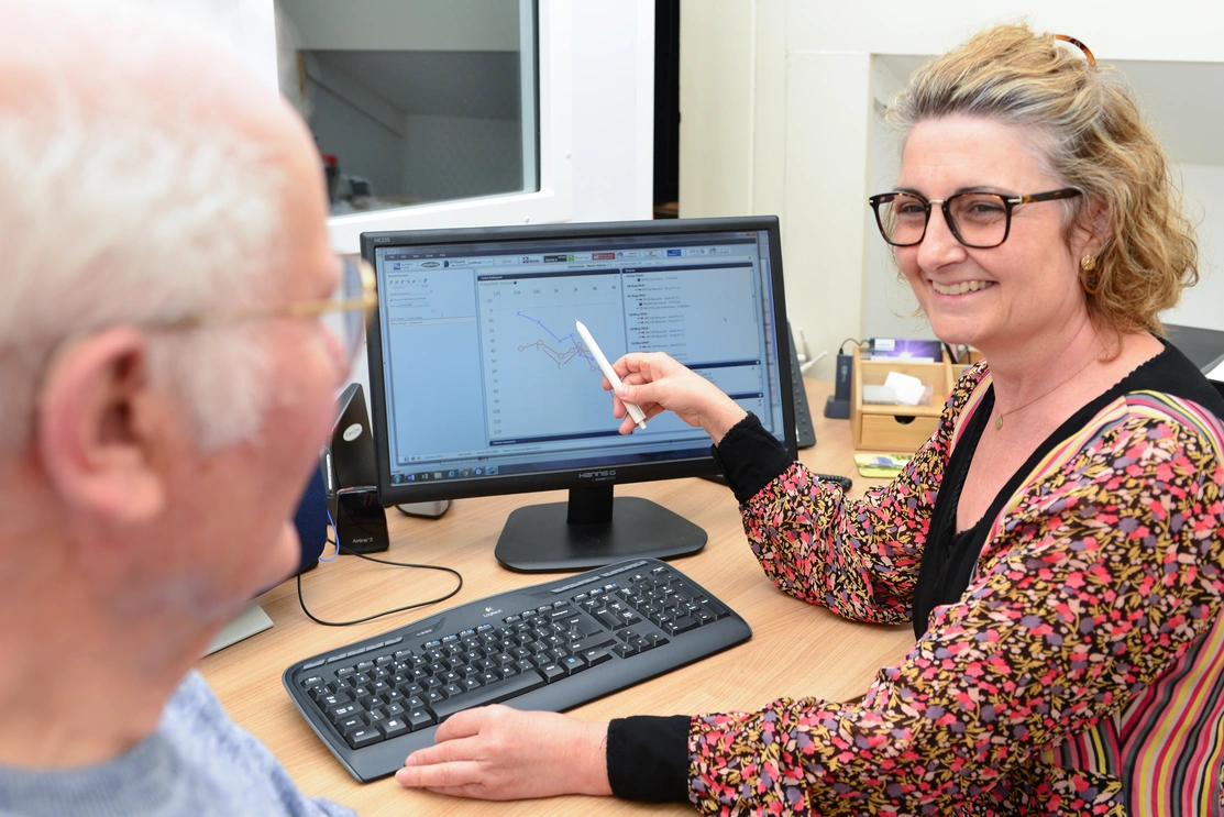 A female audiologist pointing to hearing test readings on a computer screen and smiling as an older man sat with her