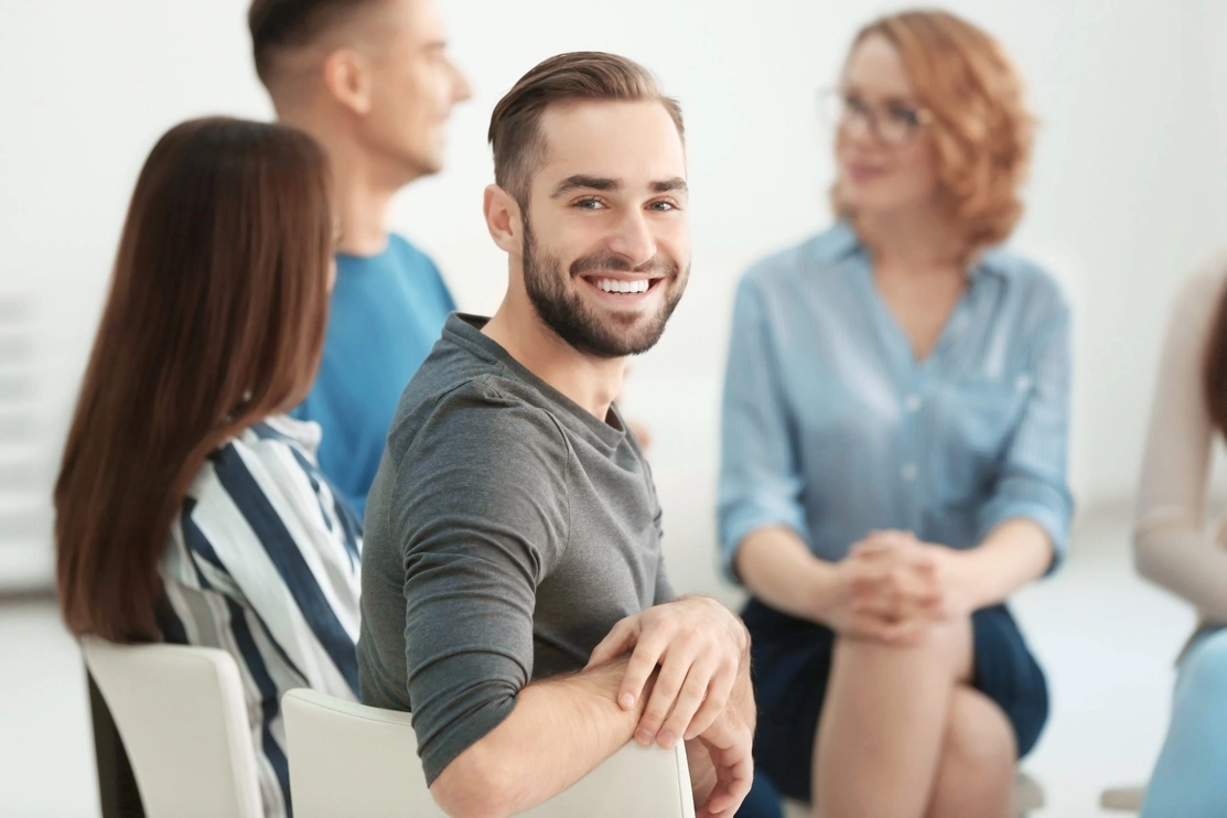 Smiling man sat in a chair turning around to look at the camera with other people sat talking in the background