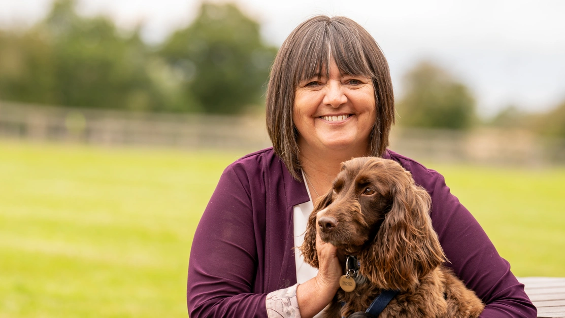 A woman sat outside smiling at the camera with a brown Spaniel sat on her lap