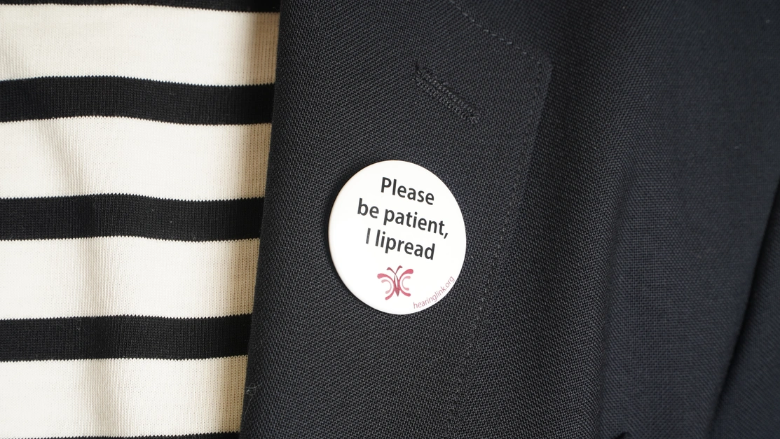 Close up of a badge on a jacket lapel that says ‘Please be patient, I lipread’