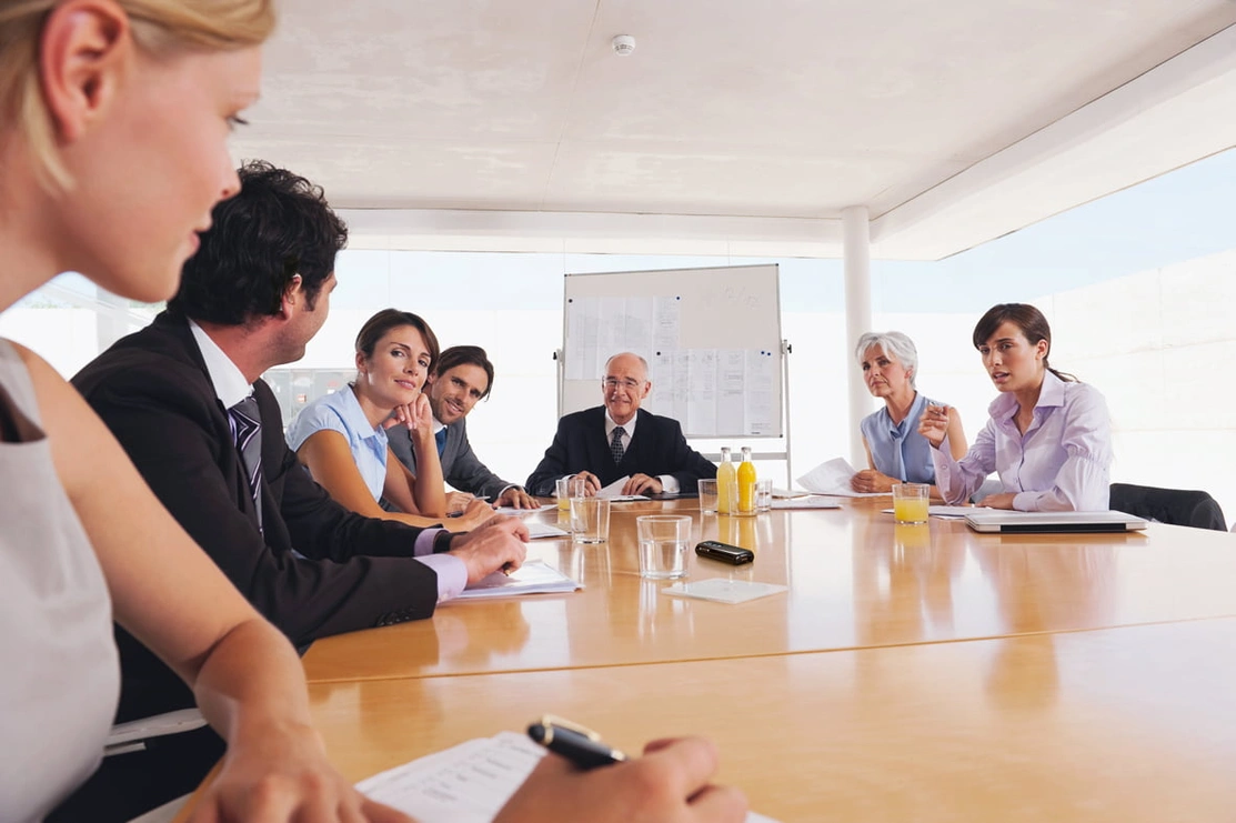 A group of people sat around a table in a work meeting talking to each other with a small black device in the centre of the table