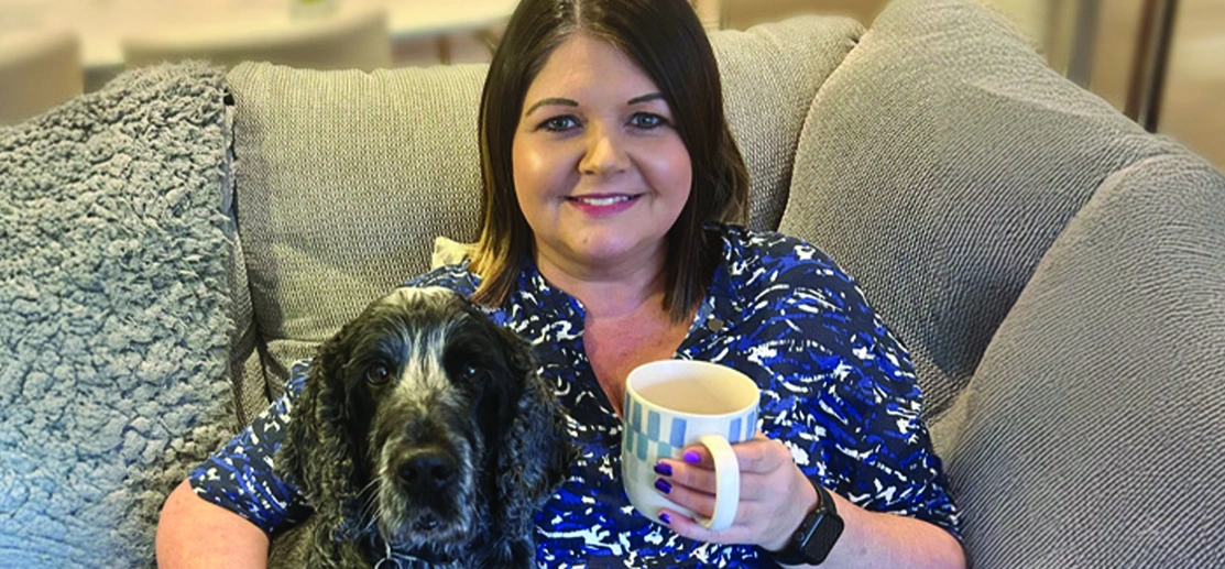 A smiling woman with brown, shoulder length hair is sitting on a sofa with her right arm is around a cocker spaniel dog