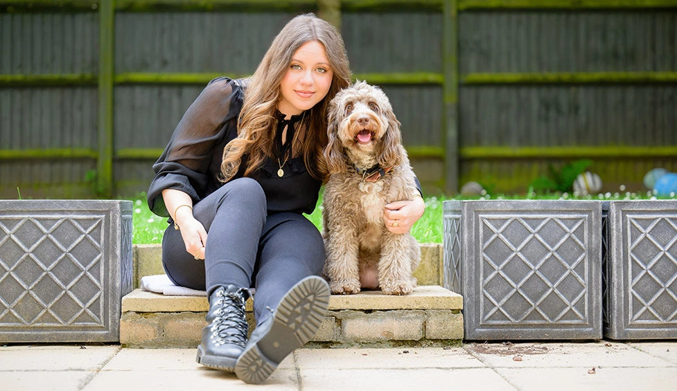 Young woman with long brown hair and dark clothes sat on a patio with her legs out in front of her and her arm around a light brown happy looking Cockapoo