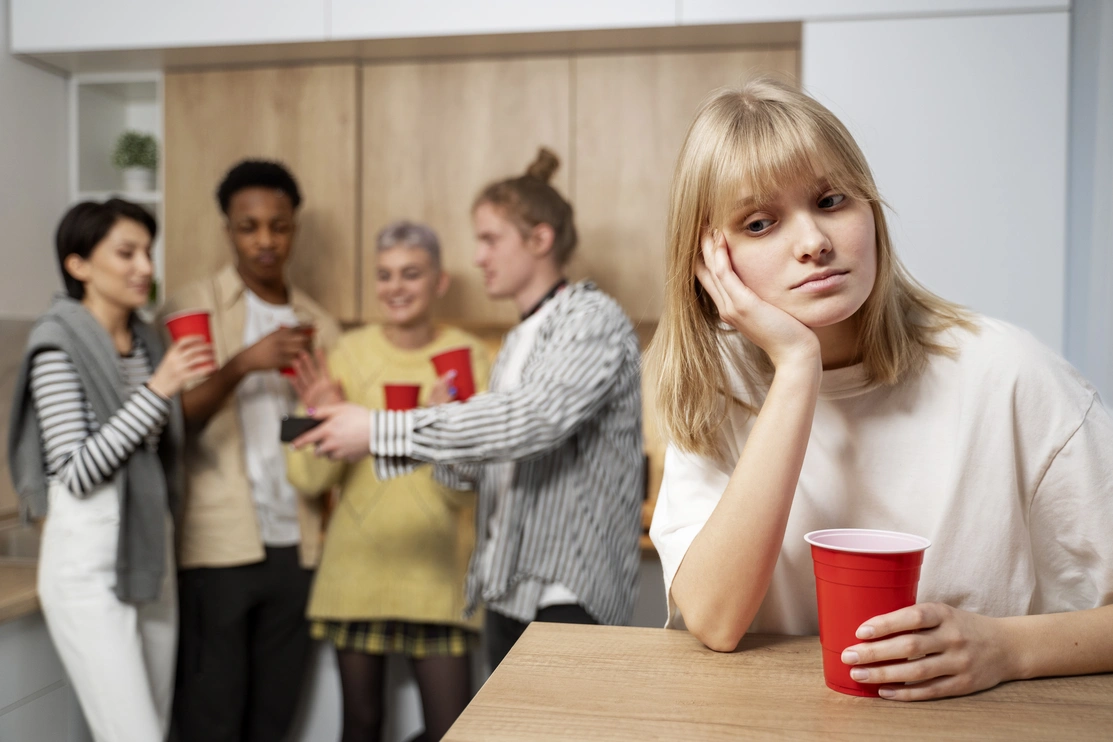 Woman in foreground sat at a table alone looking unhappy while four other people are talking happily in the background