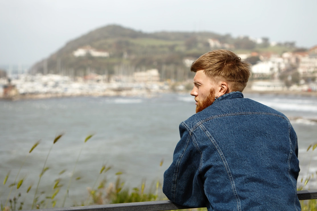 Young man looking thoughtfully out to see with boats and hillside in the distance