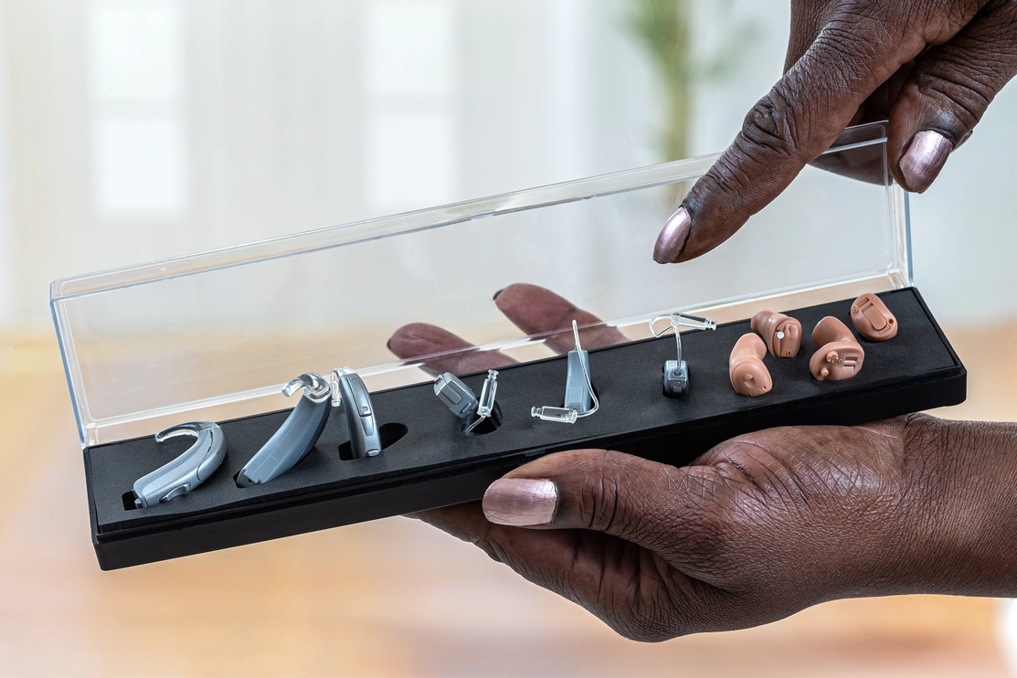 Close up of one hand holding a display case of four different types of hearing aid while the other hand points at them