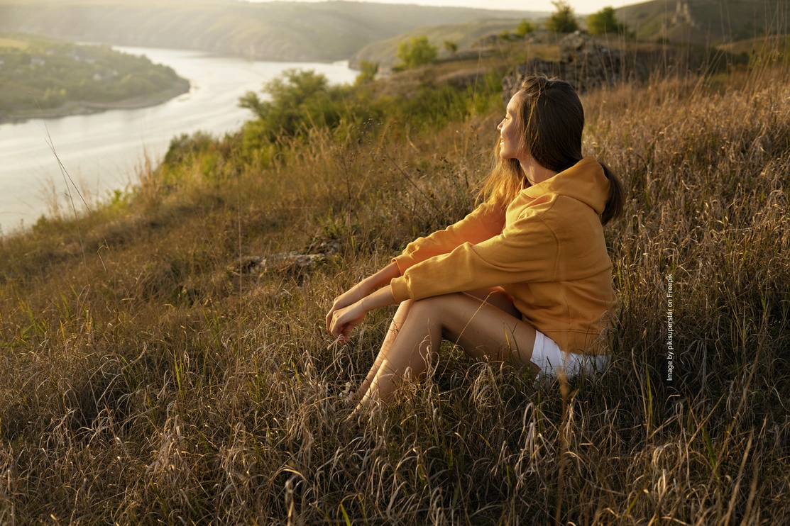 A woman sat on a hill as the sunsets looking out at some countryside