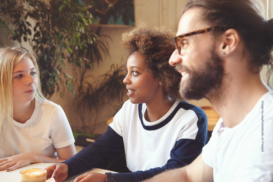 Mid shot of three people sat at a table communicating, two of them are listening and smiling as they look at the other person