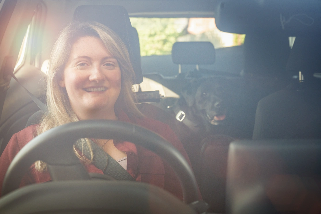 A smiing woman sitting at the steering wheel of a car. A black labrador dog is in the back seat