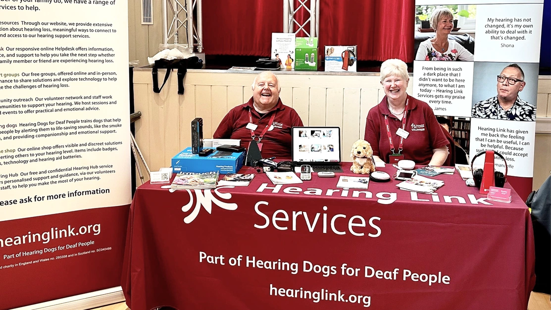 Two very happy looking people smiling at the camera, wearing hearing dogs uniform sat at a table covered in literature and clipboards