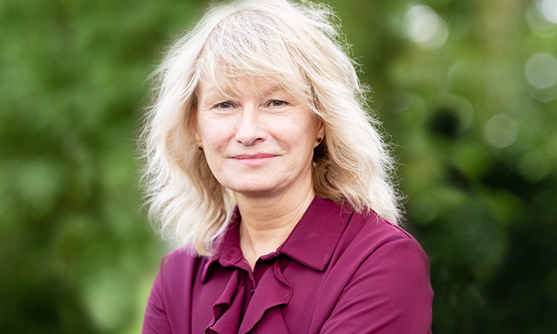 Midshot of a woman with blonde hair and a burgundy shirt sat outside with greenery in the background